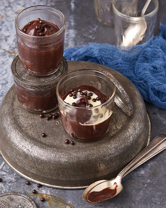 Small glass jars of chocolate pudding on top a wide upside down tarnished silver bowl next to a blue cheesecloth fabric.