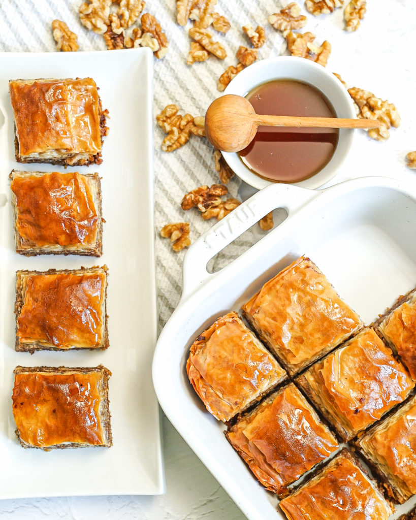 Squares of baklava in a white baking dish and a rectangular platter on a gray and white napkin with a bowl of syrup and a wooden spoon, and scattered walnuts.