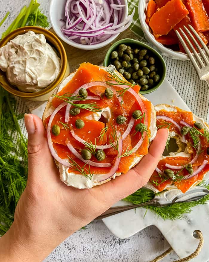 A hand holding a bagel topped with vegan cream cheese and carrot lox with capers, red onion slices, and dill, with a background of all those toppings in bowls on white table.