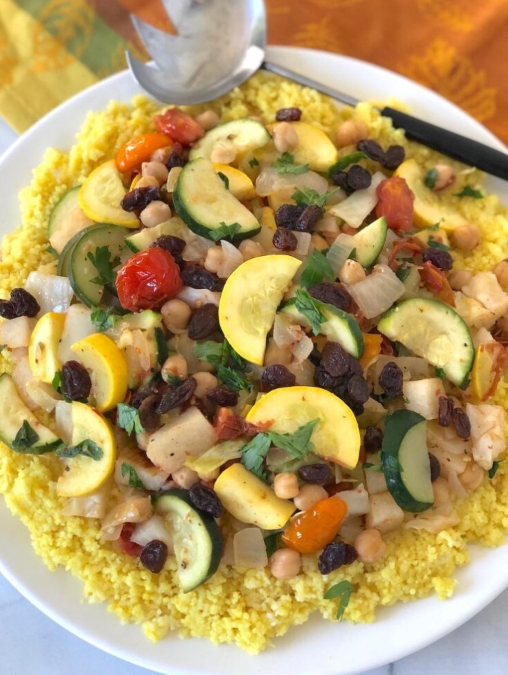 A bed of yellow couscous topped with steamed vegetables and raisins, on a multicolored table cloth with a silver and black serving spoon.