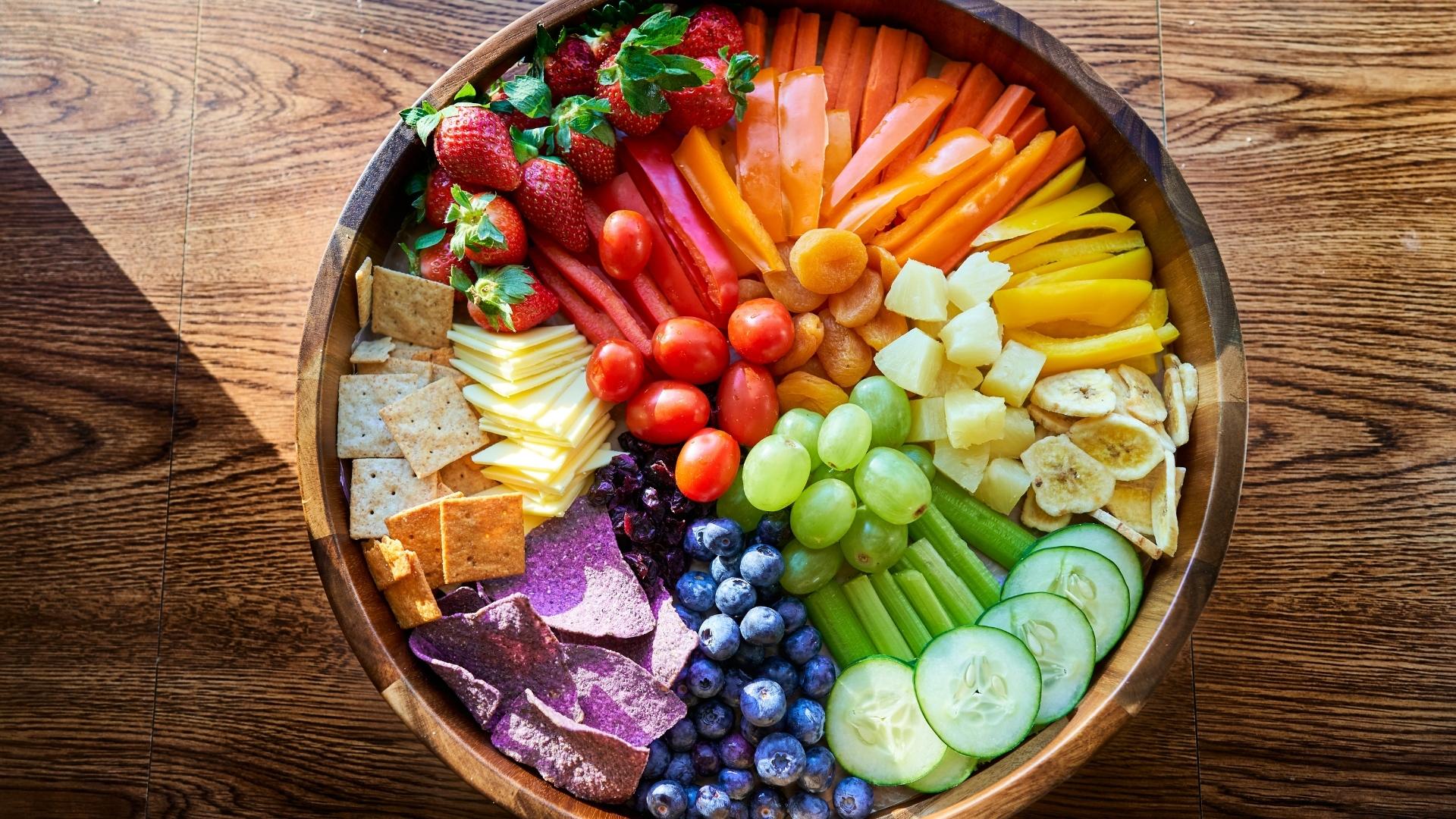 A wooden table with a large wooden bowl featuring fruit, vegetables, vegan cheese, and crackers.