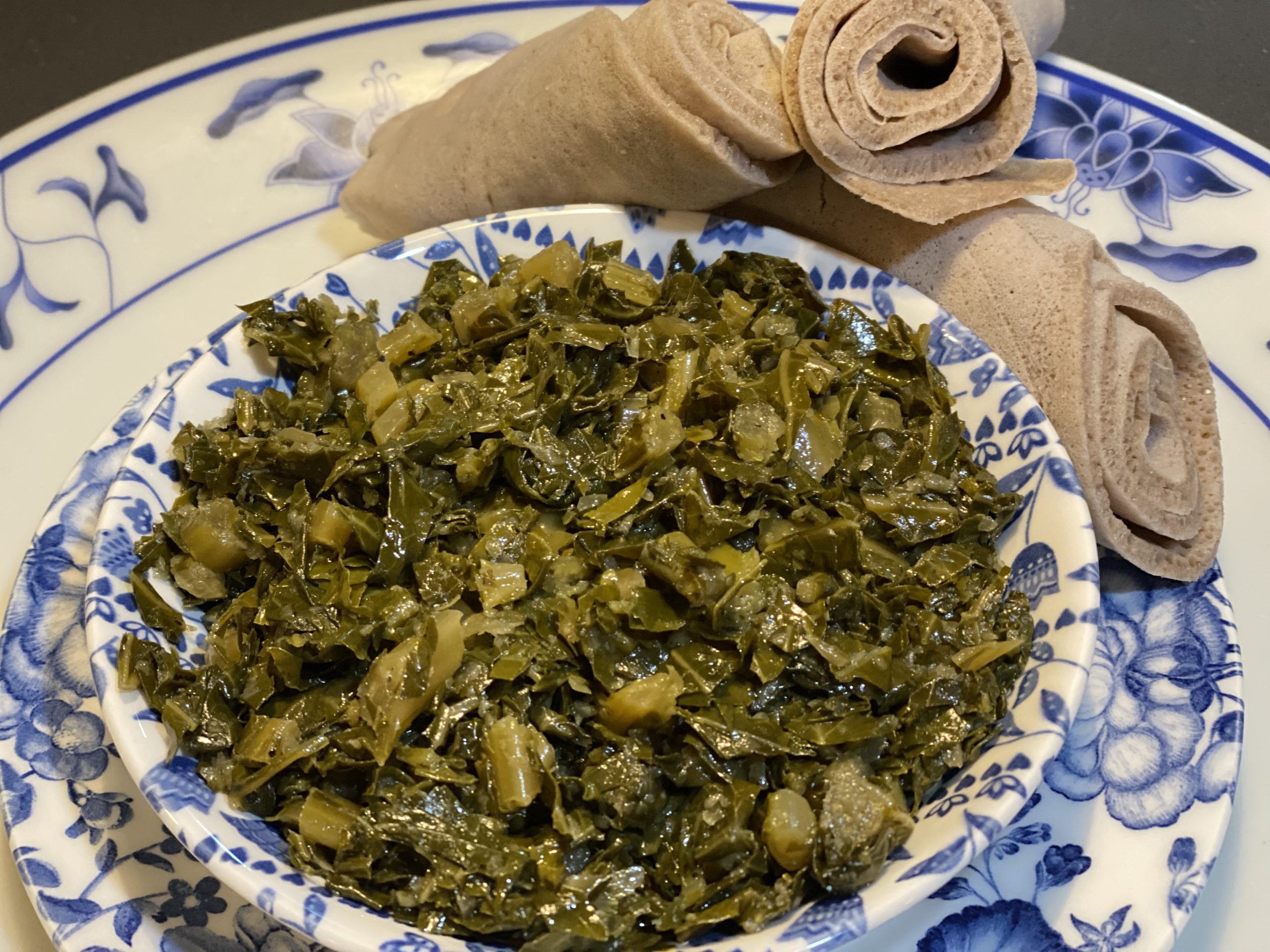 A blue and white floral bowl featuring sautéed collard greens on top of two white and blue floral plates, surrounded by three rolled up slices of injera (Ethiopian flatbread).