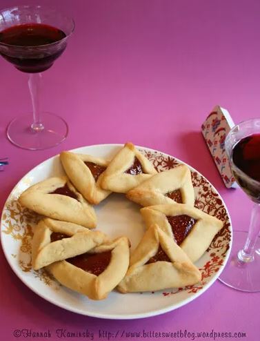A pinkish purple counter holding two glasses of wine and a red and white floral plate holding 8 fruit-filled hamentashen pastries arranged in a wreath.