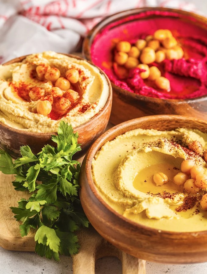Three wood bowls filled with hummus and beet hummus, garnished with olive oil, chickpeas, and paprika, all on a wooden cutting board with sprigs of parsley.