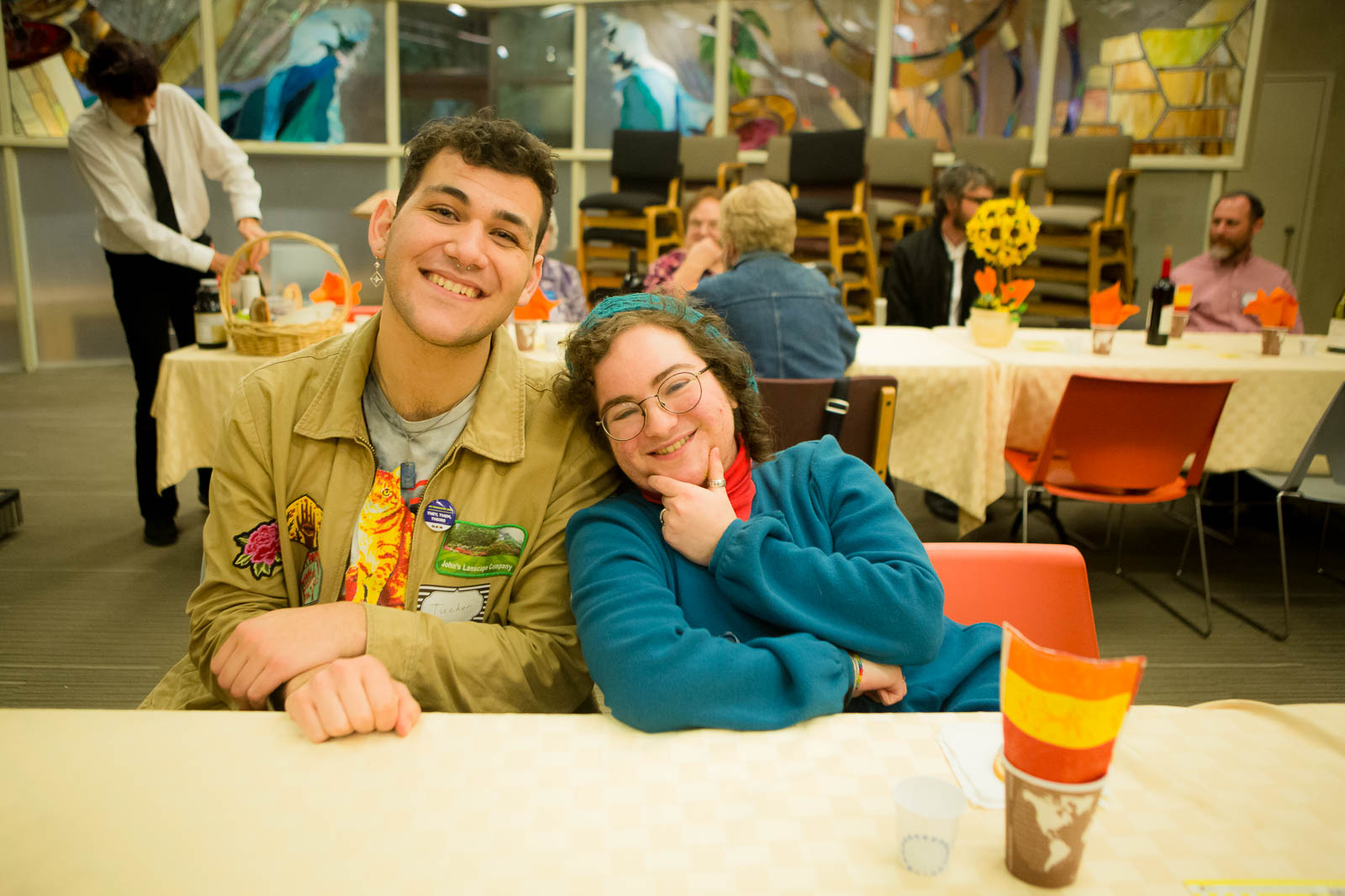 A young man and woman smiling at a table, with other tables of people in the background.