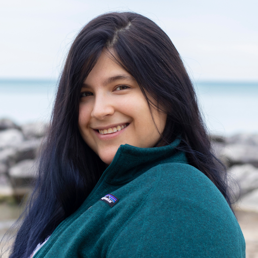 Picture of Liz Madsen standing in front of Lake Michigan with shoreline rocks in the background. They are wearing a dark green sweater.