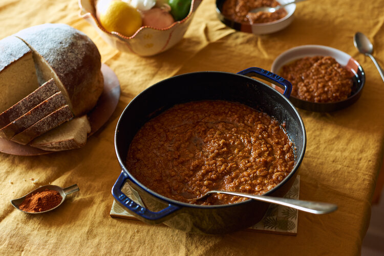 A dark blue dutch oven filled with a deep red lentil stew and a large metal serving spoon. The mustard yellow clothed table also features two bowls of the stew, a large round loaf of bread with several slices cut, a fruit bowl, and a little leaf-shaped plate of berbere spice mixture.