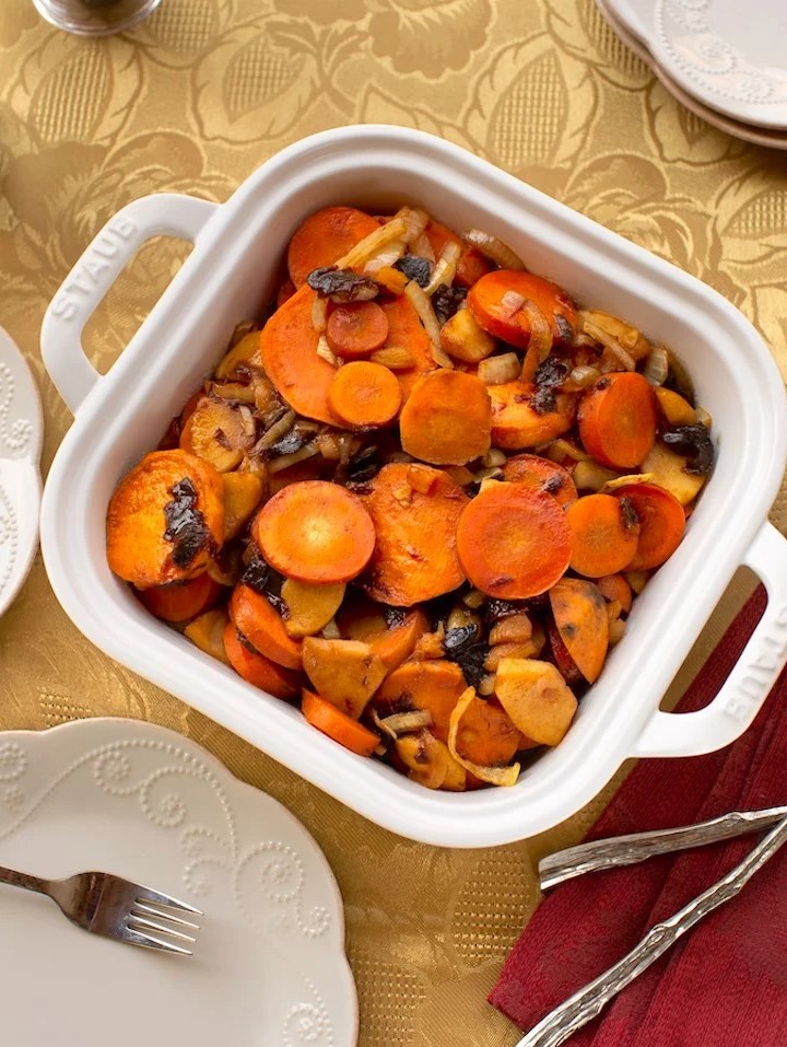 A white ceramic baking dish filled with cooked carrots, sweet potatoes, and dried fruit on a gold floral tablecloth surrounded by white plates and silver forks.