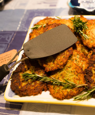 A square white plate filled with latkes and sprigs of rosemary, and a black spatula.