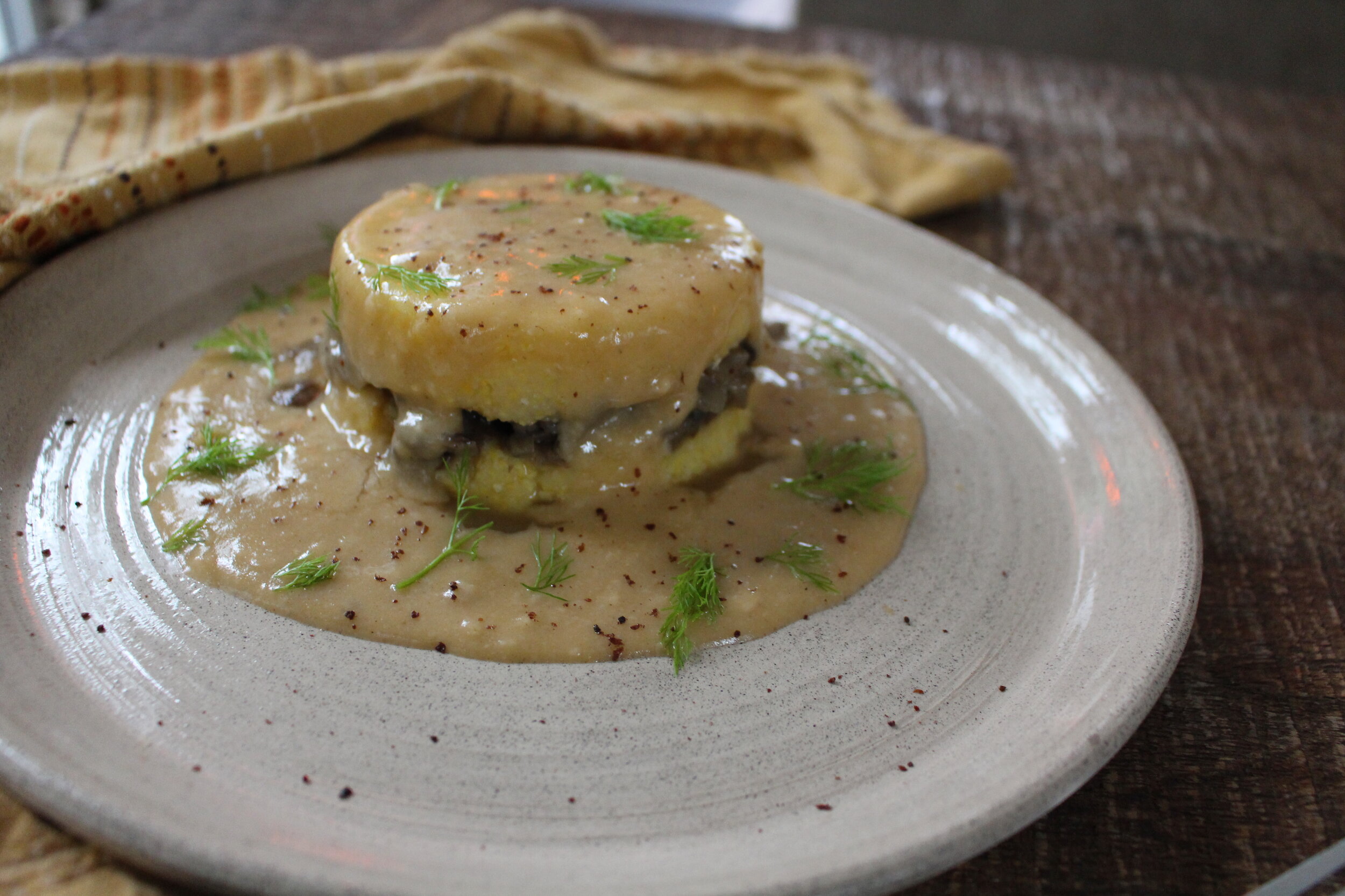 Two patties of polenta with a layer of cooked mushrooms in between and drenched in pale gravy with dill pieces and pepper on a white plate with a yellow napkin on the side.