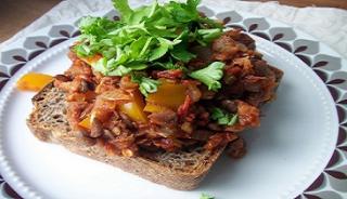 A dark piece of toast topped with a sloppy joe mixture and lots of fresh herbs on a white plate with a gray and purple leaf design.