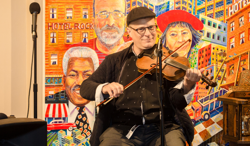A seated man playing the violin and smiling for the camera in front of a vibrant painting featuring people and buildings.