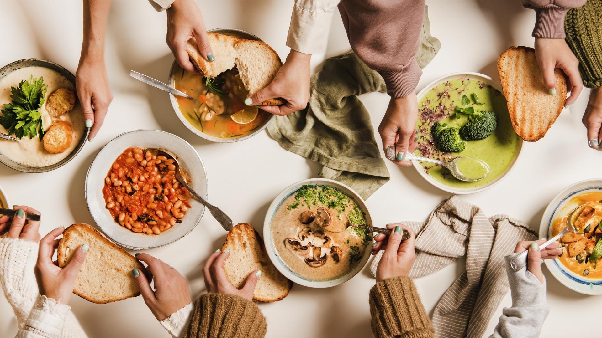 A banner of various bowls of plant-based food on a table with many different hands holding food and slices of bread.