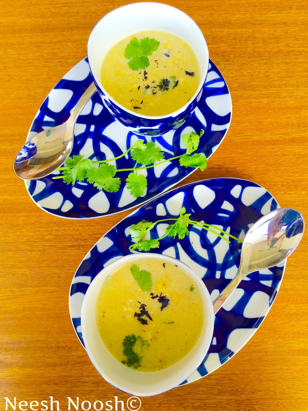 Blue and white abstract plates with small bowls of yellow soup and cilantro all on an orangish wood table.