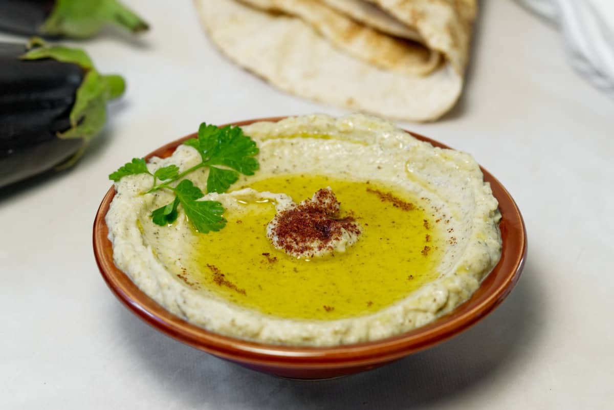 A red bowl filled with baba ganoush, topped with olive oil, paprika, and parsley on a white table with flatbread and eggplant in the background.