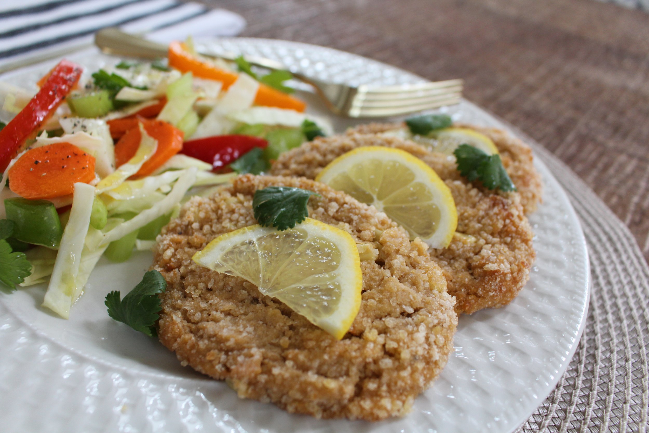 A white plate with corn schnitzel with lemon slices, parsley, and a side salad all on a tan placemat on wood table.