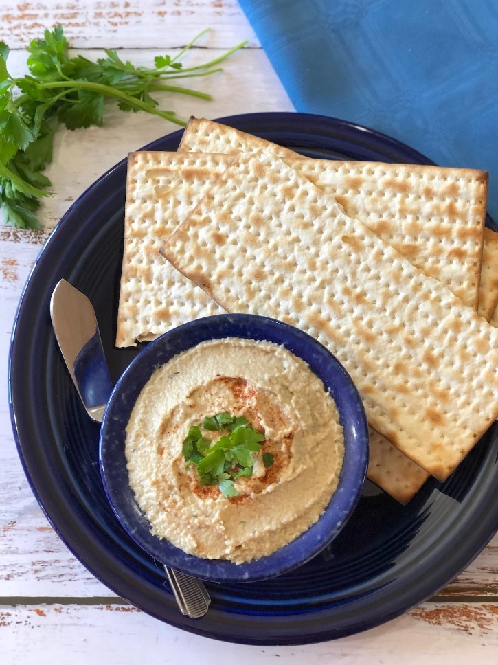 A dark blue plate featuring several pieces of matzah, a butter knife, and a small dark blue bowl of a light brown paste. Surrounded by a blue napkin and bunch of parsley on a white wood table.