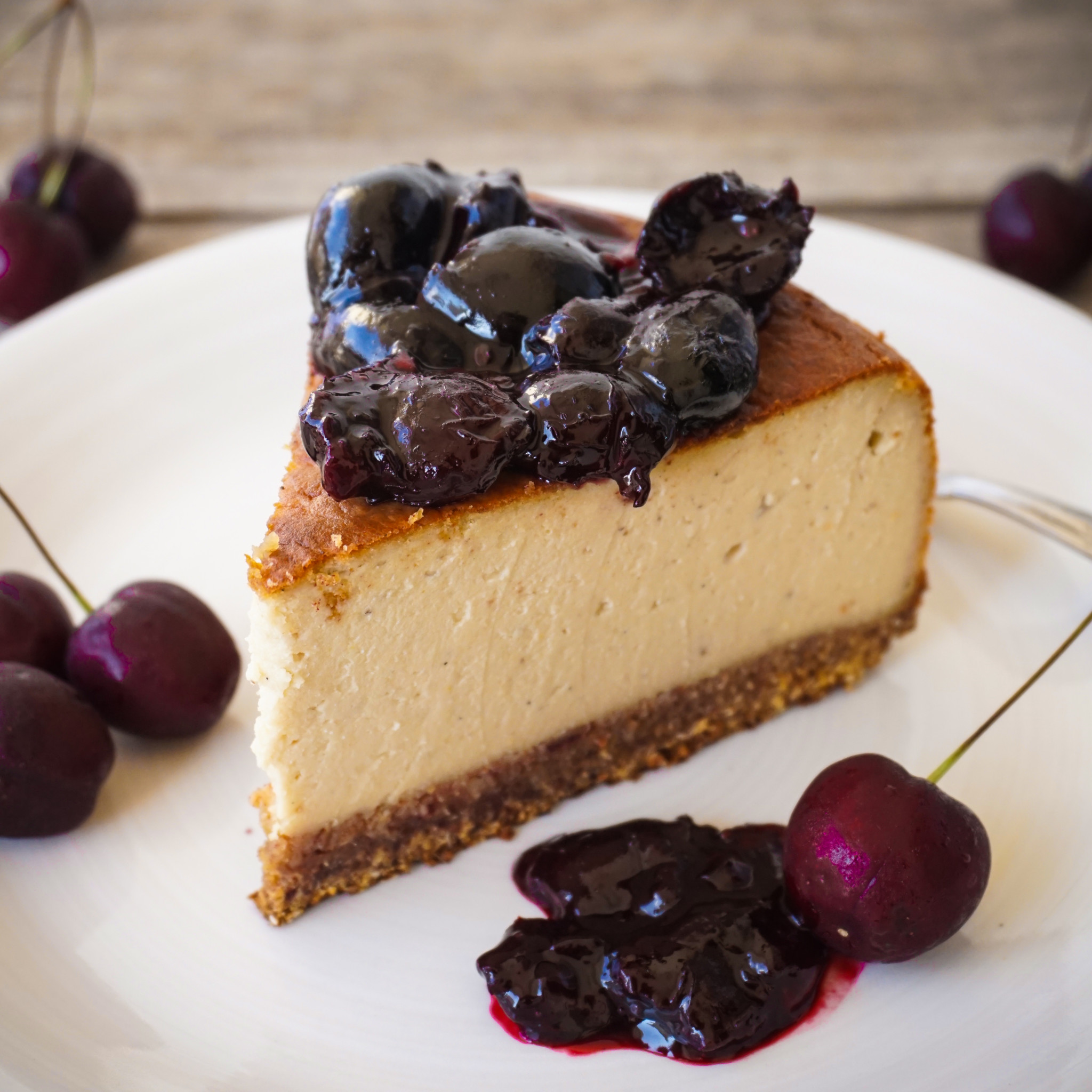 A slice of cheesecake, topped with cooked black cherries on a white plate with fresh black cherries in the background.