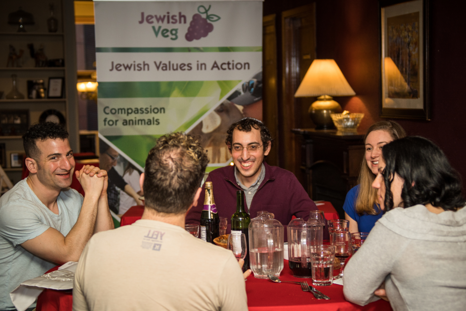 A group of young adults sitting at a red clothed table with food and wine, a banner for Jewish Veg hanging in the background.