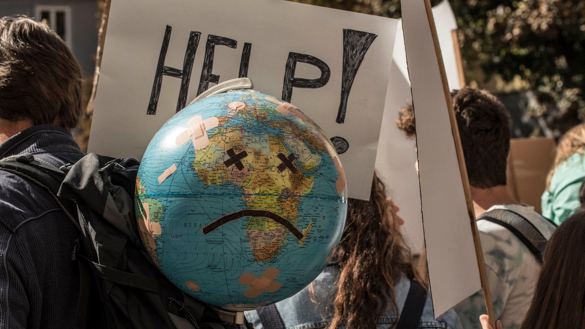A closeup of signs in a public protest: a white sign that says HELP! in black marker, and a globe with a frown face and bandaids taped on it.