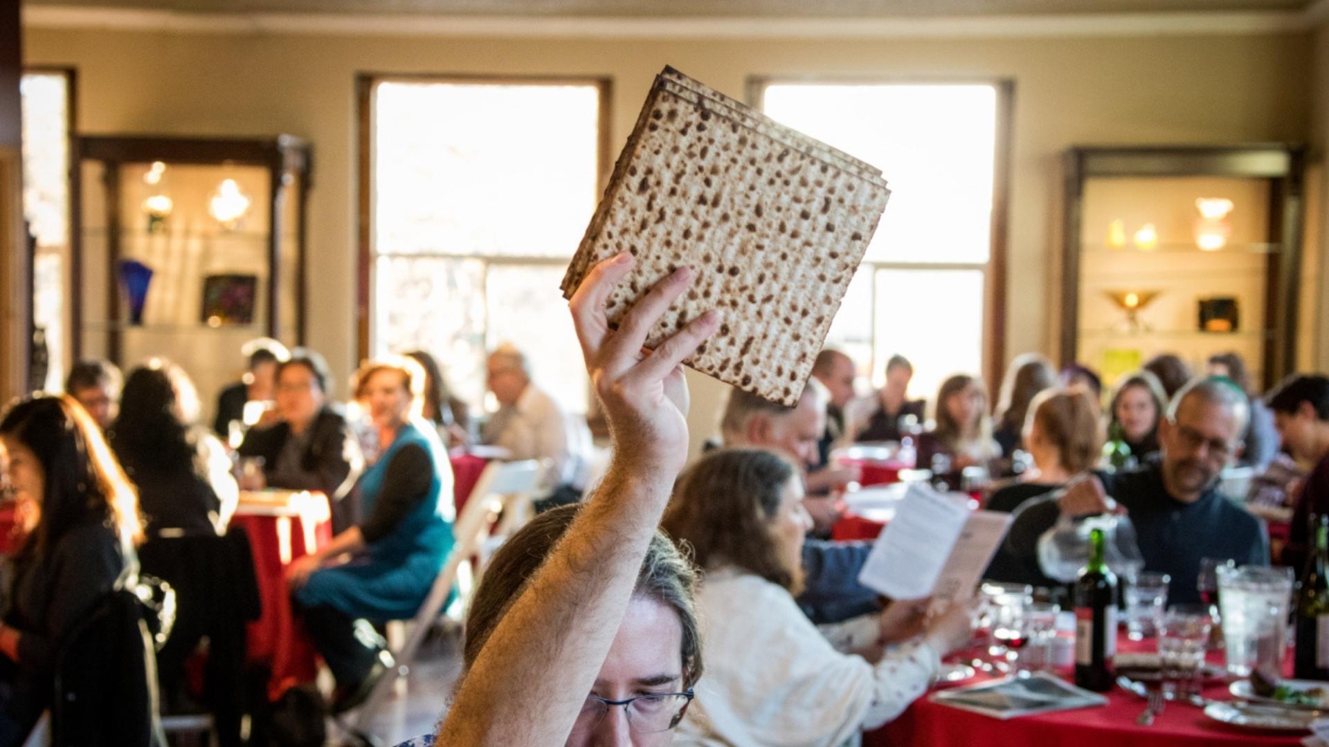 A man holding up four squares of matzah in a crowded room filled with people sitting at round tables.