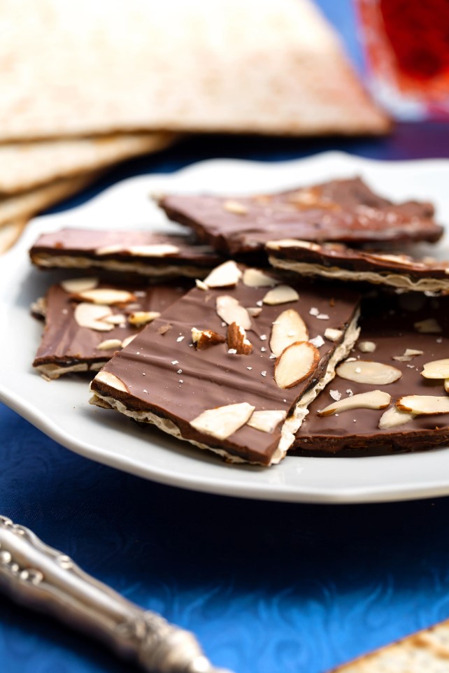 A white plate with squares of chocolate and nut covered matzah on a blue placemat, whole squares of matzah in the background.