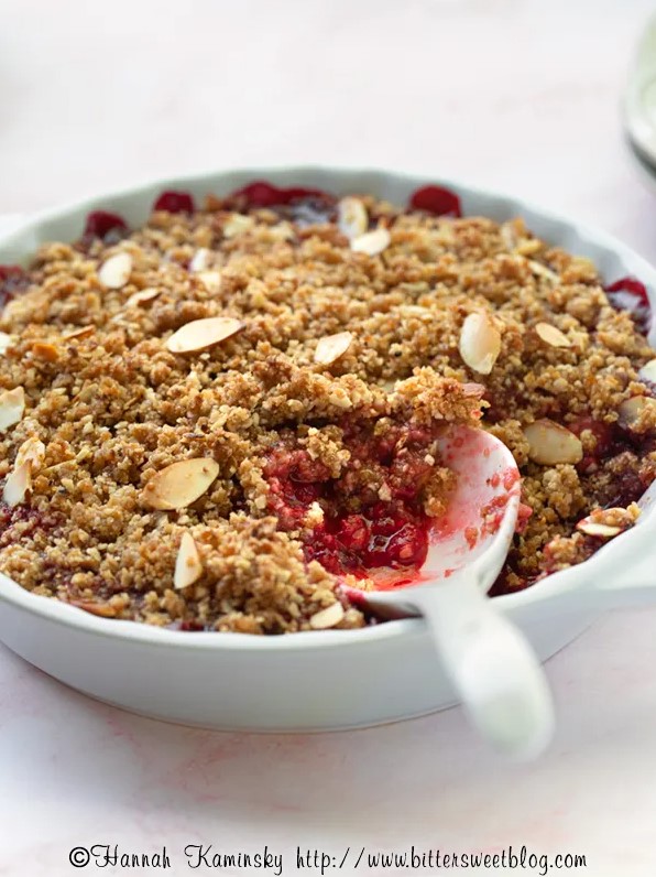 A white bowl filled withe cooked red berries topped with a nutty crumble and a white spoon dug in.