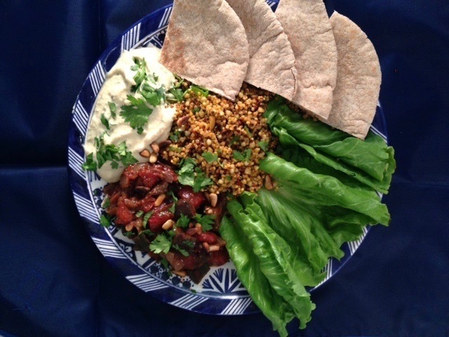 A blue and white plate topped with hummus, cooked eggplant salad, grains, greens, and pita wedges, all on a navy tablecloth.