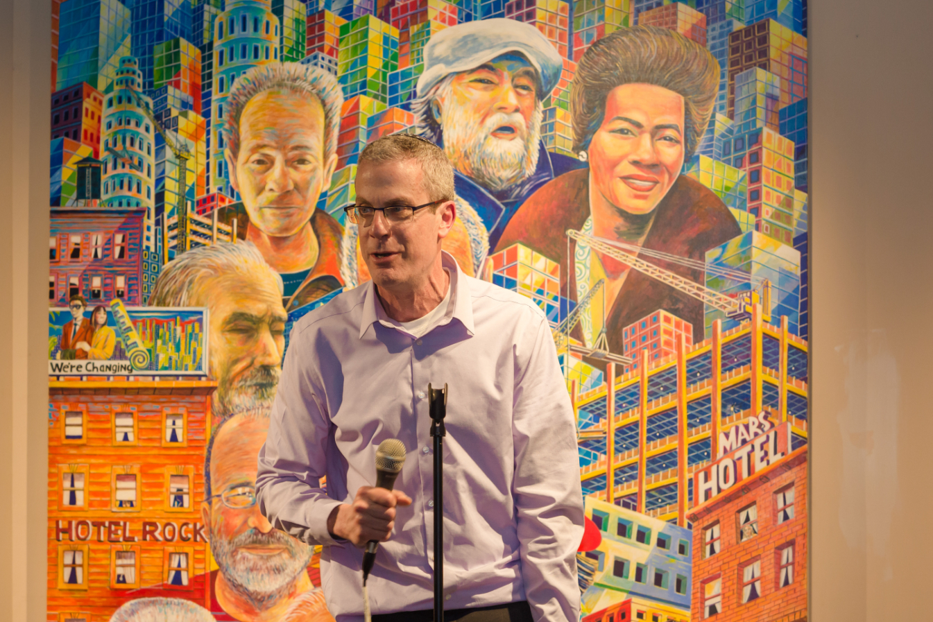 Executive Director Jeffrey Spitz Cohan speaking with a microphone in front of a vibrant painting featuring people and buildings.