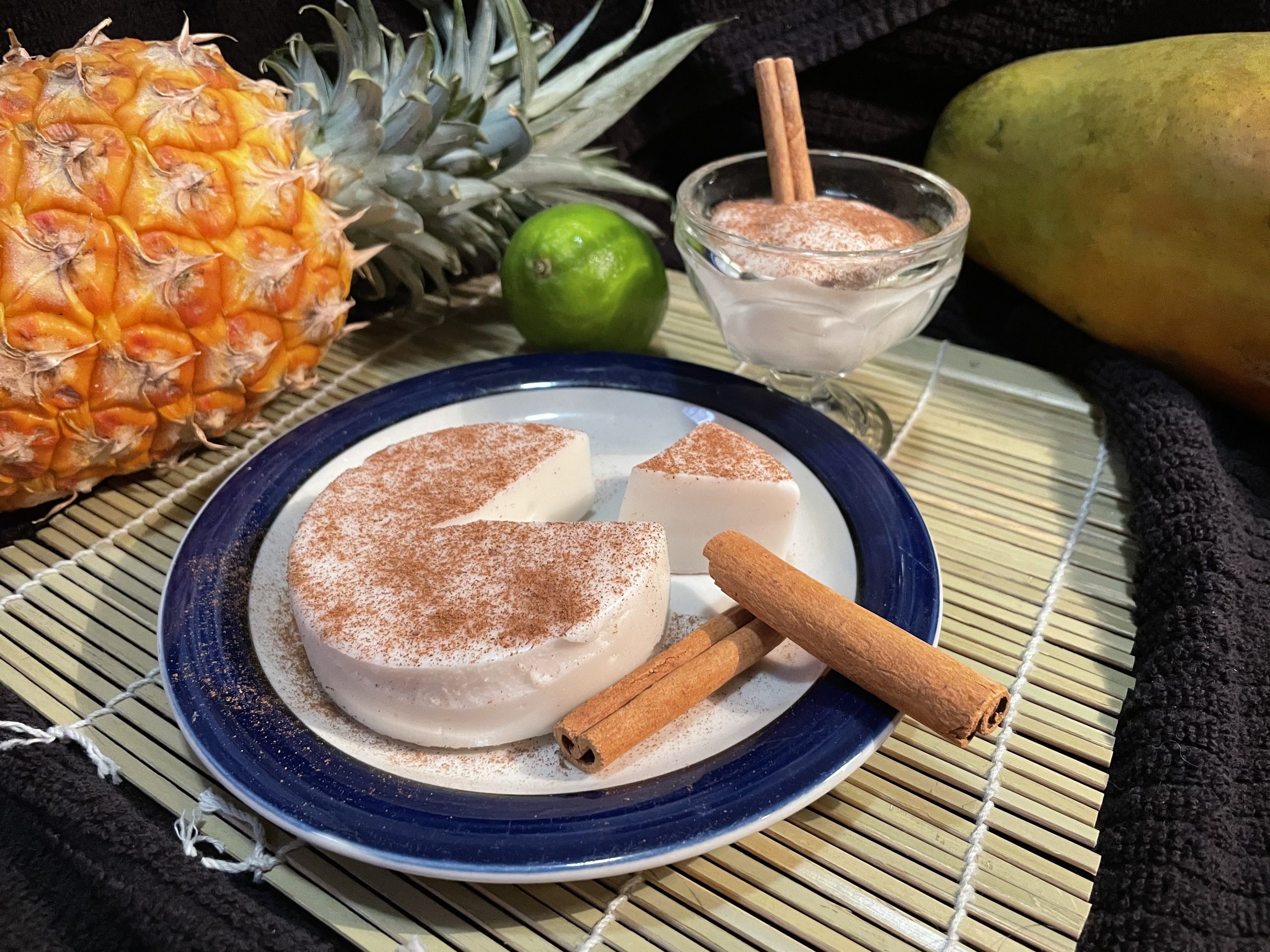 A blue rimmed plate featuring a round tembleque, which is a solid custard topped with cinnamon. Surrounded by cinnamon sticks, papaya, pineapple, lime, and a custard dish filled with more custard all on a bamboo mat and black kitchen towel.