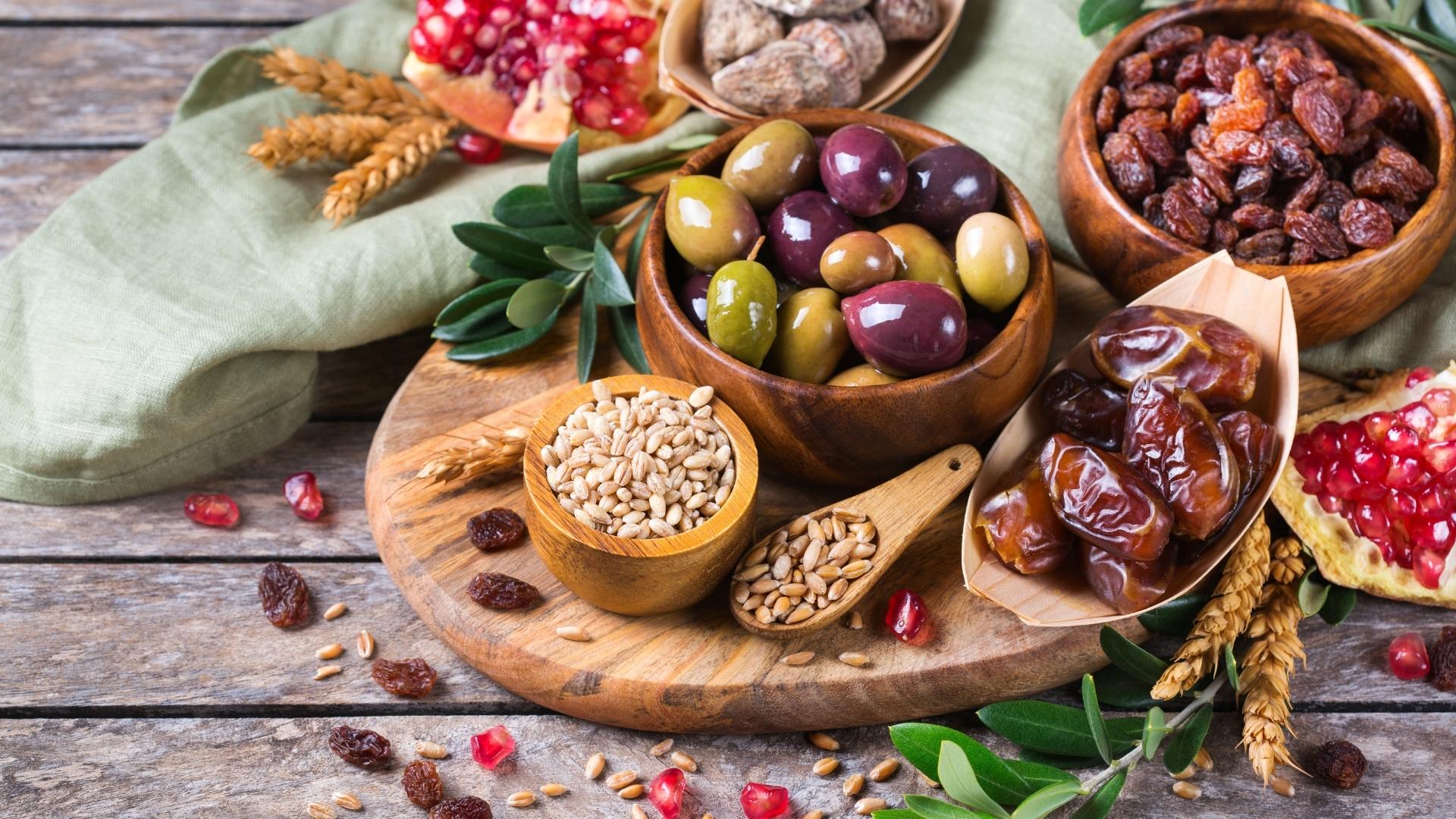A group of wood bowls filled with olives, dates, grains, pomegranate arils, and raisins, surrounded by a green napkin on a brown wooden table.
