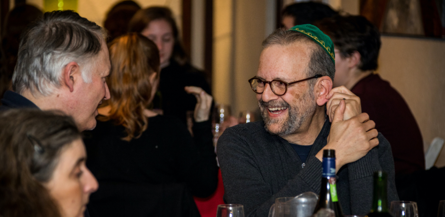 A man in a green yarmulke / kippah and black shirt laughs while conversing with another man, surrounded by other people in the background.