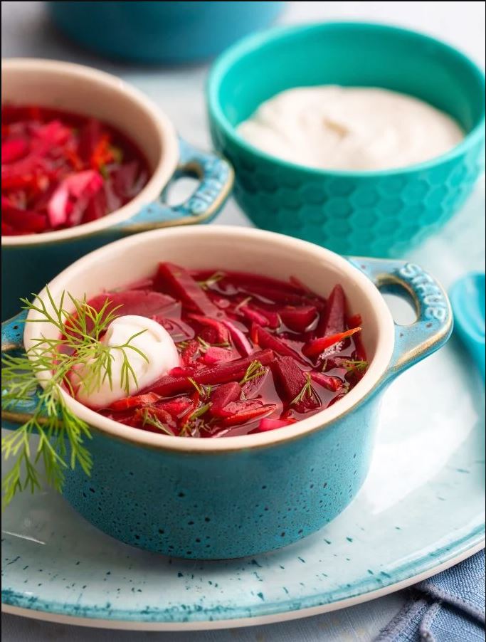 Bright turquoise coquettes filled with bright red chunky borscht soup, garnished with a dollop of vegan sour cream and a sprig of dill.