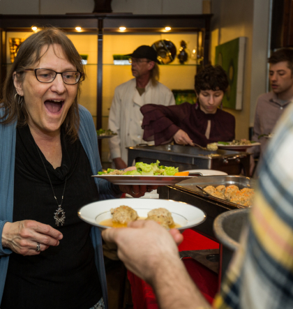 A woman very excited to be receiving a bowl of matzah ball soup someone is handing her.