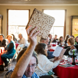 A man holding up four squares of matzah in a crowded room filled with people sitting at round tables.