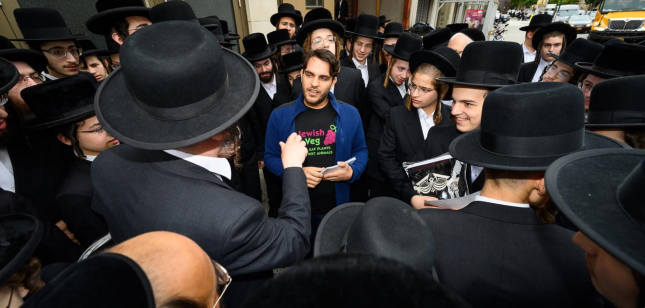 A male volunteer wearing a Jewish Veg shirt, is surrounded by attentive Chassidic men on a busy city street.