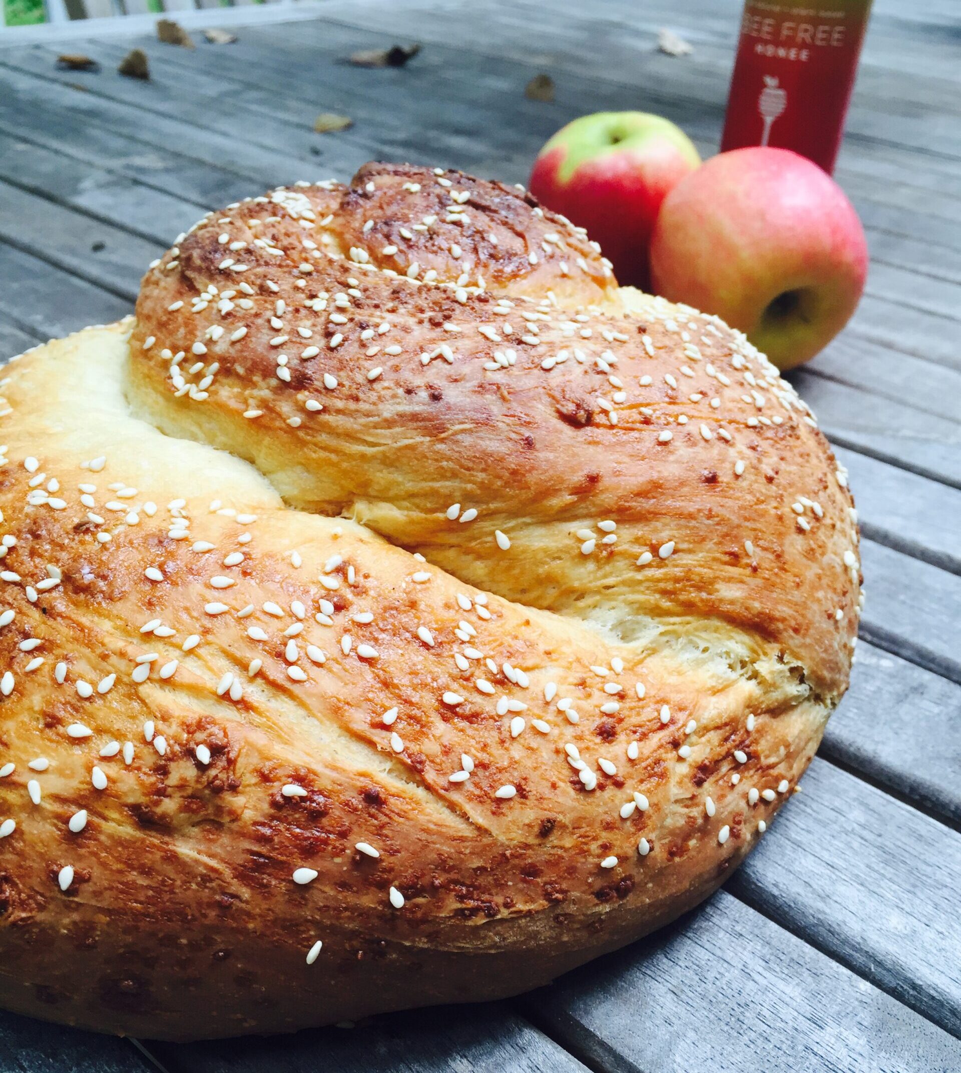 A closeup of a round vegan challah covered in sesame seeds, with apples and bee free honee in the background.