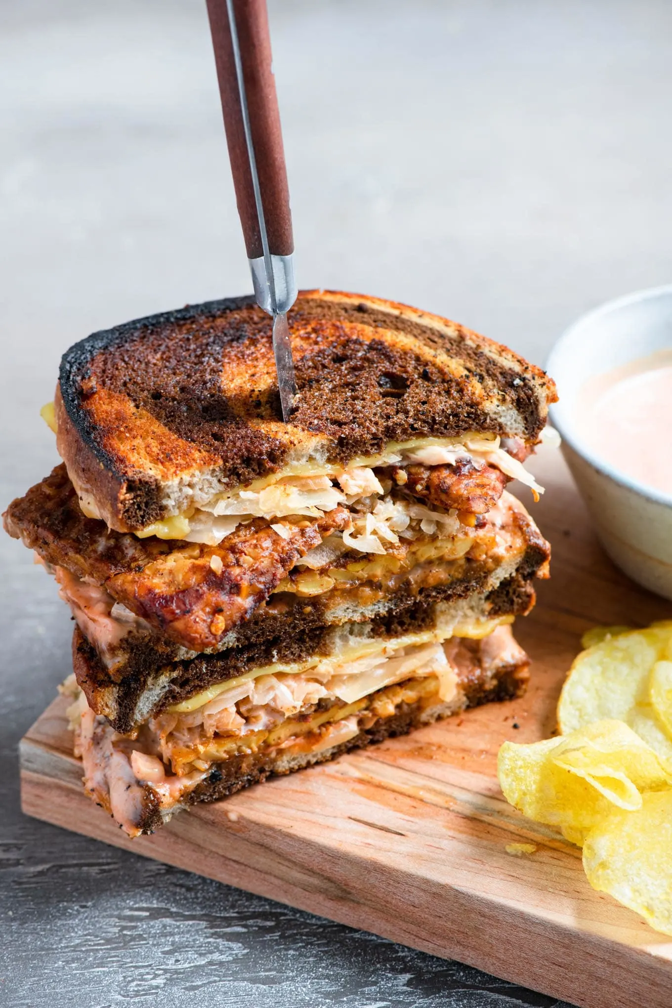 A wooden board featuring a sliced reuben sandwich stacked with a steak knife stuck in them, a bowl of light orange sauce and potato chips on the side.